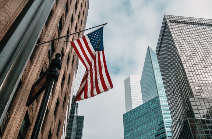the USA or United States of America flag on a flagpole near skyscrapers under a cloudy sky Fiemg atrai interesse dos Estados Unidos e Reino Unido para cooperação em terras raras e minerais críticos. O Brasil, com 23% das reservas mundiais, é chave na transição energética e tecnologias de defesa.